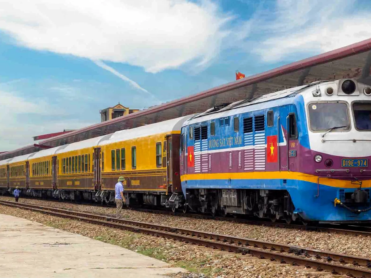 A blue diesel locomotive and yellow carriages beside a station platform under a blue sky, with a railway worker walking along the track