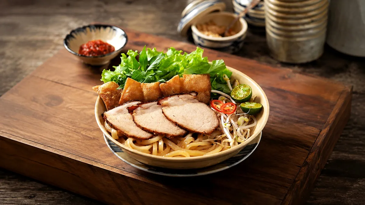 A bowl of cao lầu with sliced char siu pork, crisp noodle pieces, herbs and chili