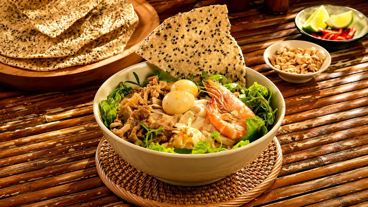 A bowl of mì Quảng with shrimp, pork and quail eggs and a sesame rice cracker on top