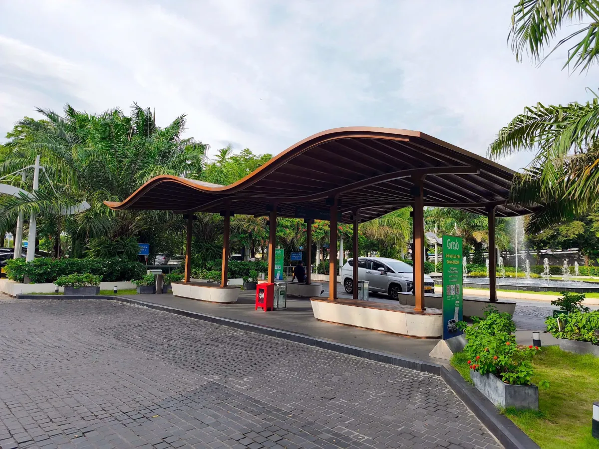 A covered waiting shelter with a wavy wooden roof and stone benches in the airport car park, with a green Grab sign marking a GrabCar pickup zone