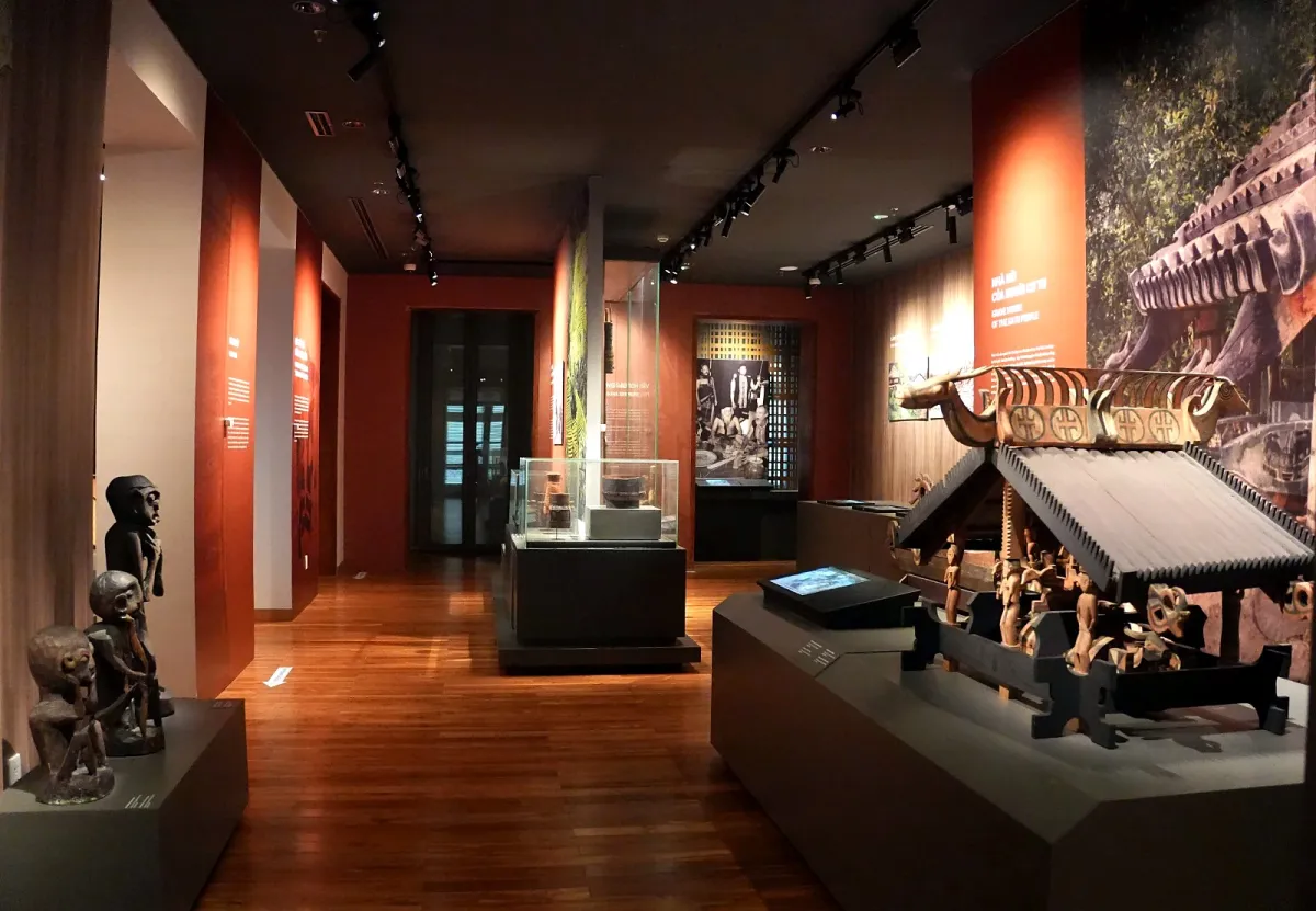 A dimly lit museum gallery with carved wooden statues and a Katu grave house on a platform