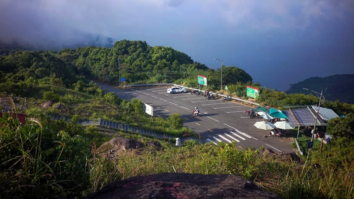 A hillside parking area with a taxi, parked motorbikes and two people riding a motorbike, with the sea and low cloud behind