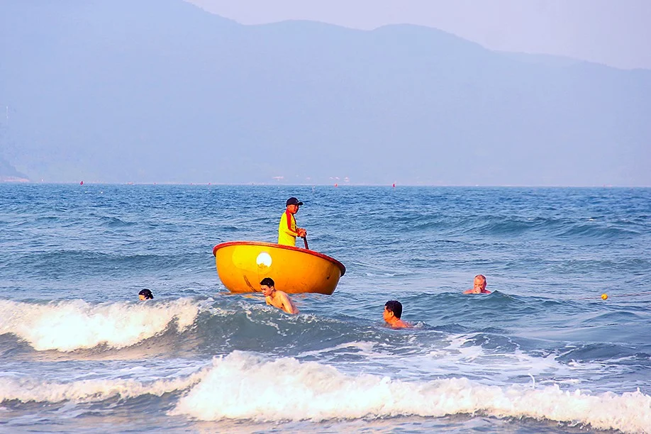 A lifeguard in a yellow and red uniform standing in a yellow basket boat among swimmers in the waves, with mountains behind