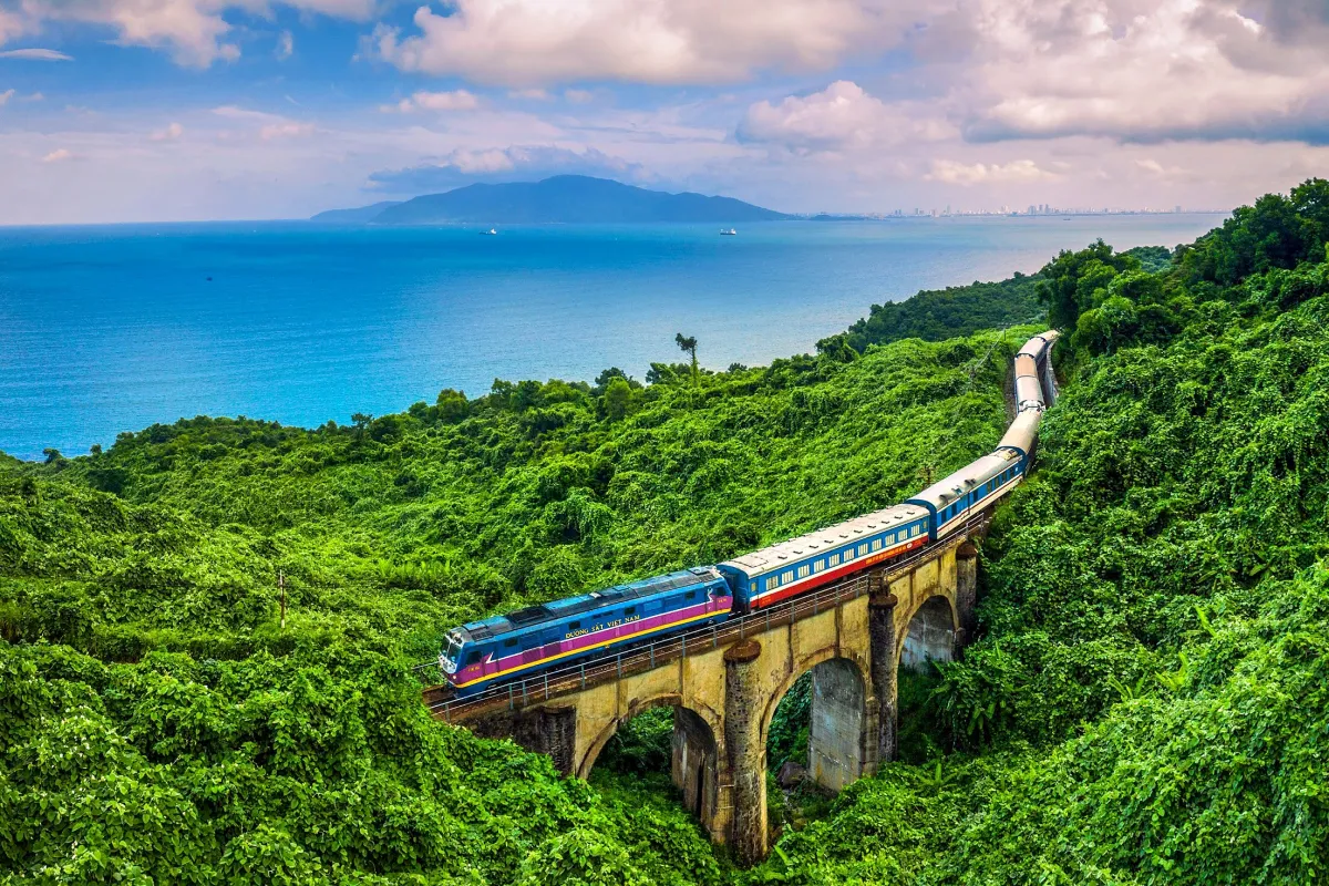 A passenger train crossing an arched stone viaduct through green hills above a blue bay