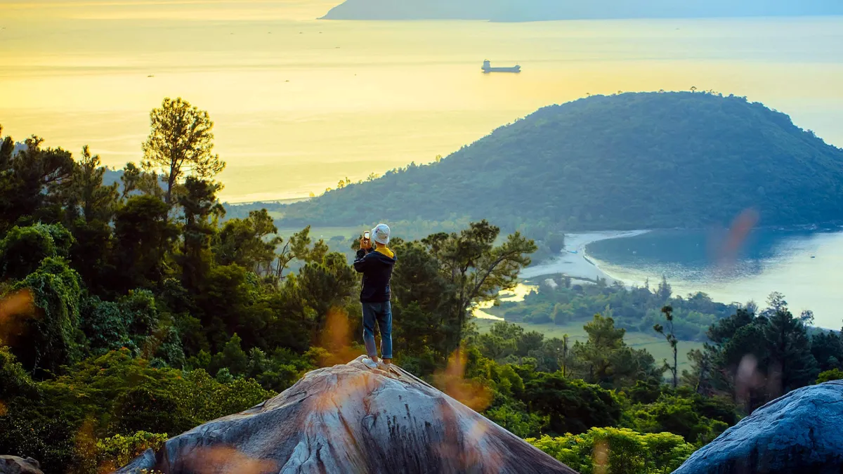 A person standing on a granite boulder and photographing a bay and a green peninsula in golden light