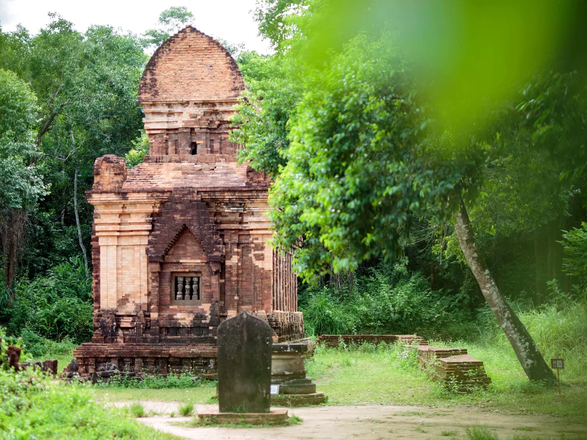 A red-brick Cham tower with a stone stele in front, surrounded by green trees at My Son