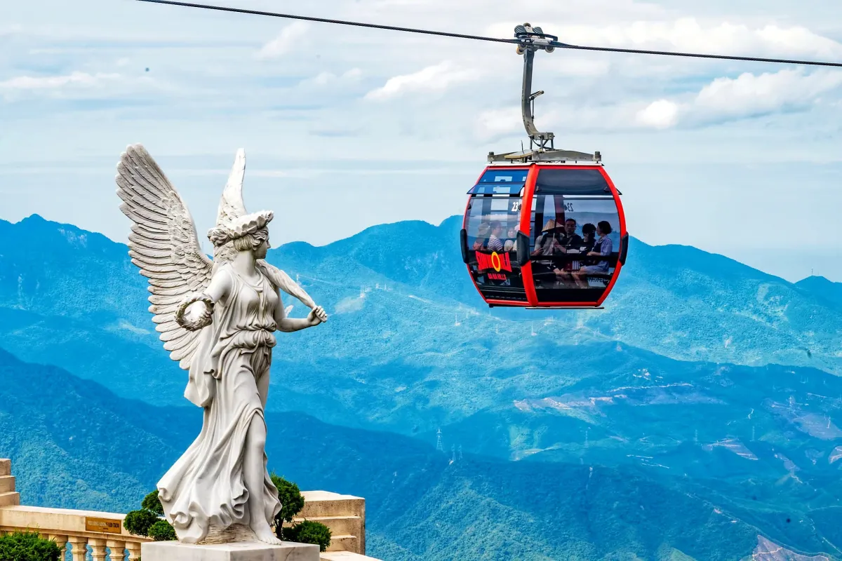 A red cable car cabin passing a white angel statue above green mountains at Ba Na Hills