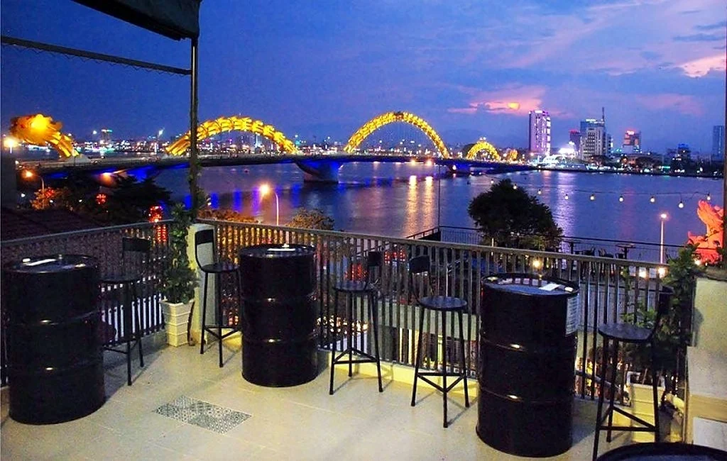 A riverside terrace with barrel tables and high stools, looking across the Han River at Dragon Bridge lit yellow at dusk