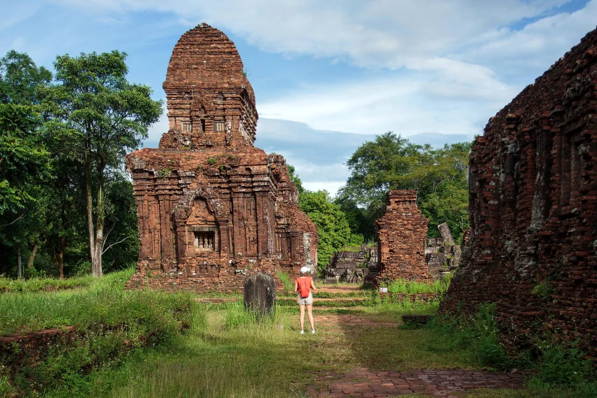 A tall red-brick tower among green grass and trees at My Son, with a visitor seen from behind on the path