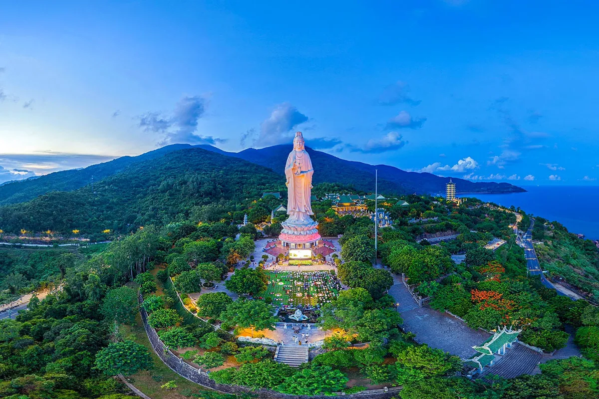 A tall white statue on a lit lotus base above a pagoda courtyard, with green hills and the sea at dusk