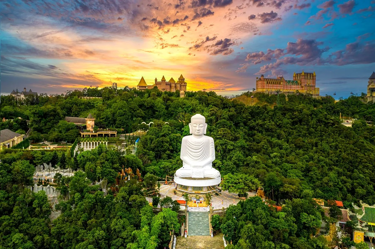 A white Buddha statue among the trees at Ba Na Hills, with the French Village behind at sunset