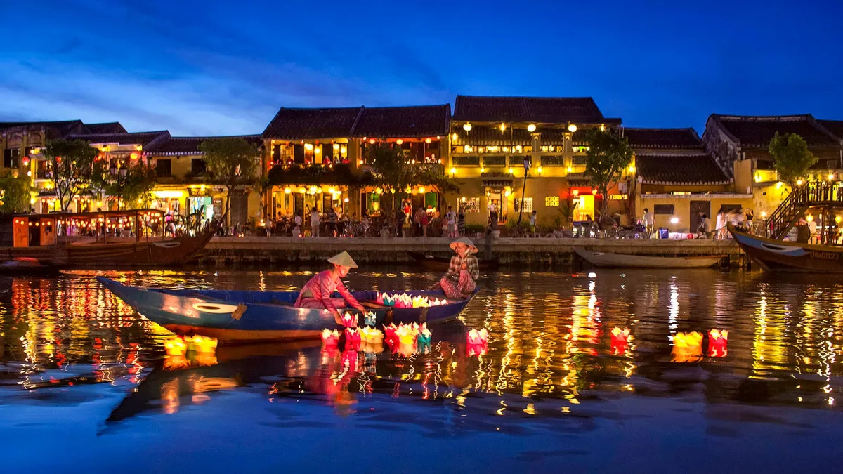 A wooden boat on the Hoai River at dusk, with two rowers setting small lanterns on the water