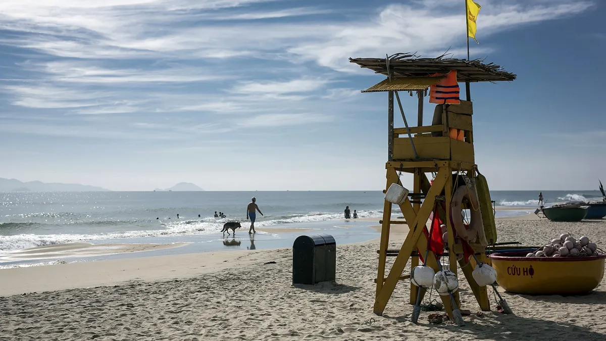 A wooden lifeguard chair with a thatched roof, life rings and a yellow flag on An Bang beach, a yellow basket boat marked Cuu ho beside it and swimmers in the waves