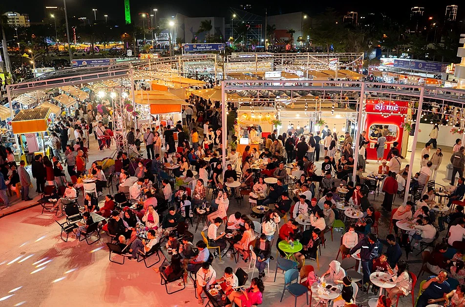 Aerial view of a dense evening crowd at tables between lit food stalls at B.Fair, with a red Si:mê stall on the right