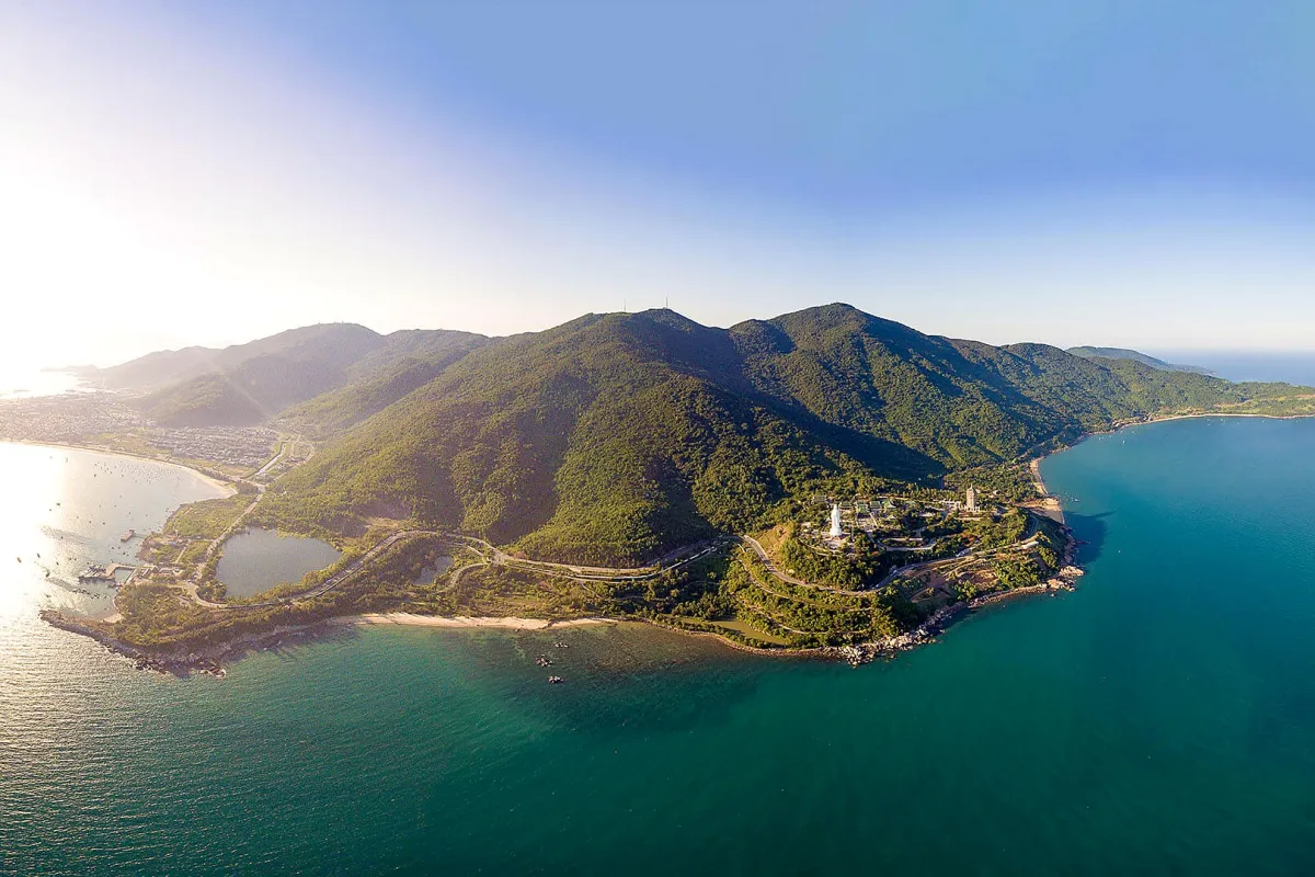 Aerial view of a green mountain peninsula with a road along the coast, a white statue on the hillside and the city across the bay