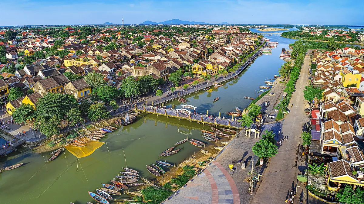 Aerial view of Hoi An with the Hoai River in the middle, tiled roofs of the old town on the left, a wide riverside street on the right and a footbridge with two white arches