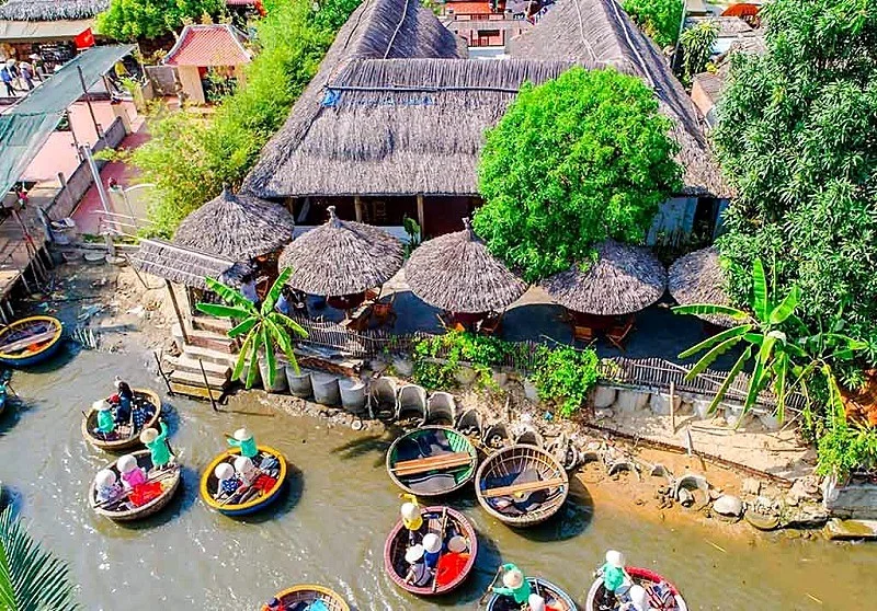 Aerial view of round basket boats with passengers in conical hats along a riverbank beside a building with thatched roofs and thatched parasols