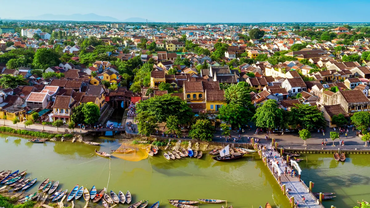 Aerial view of the Hoai River in Hoi An with moored wooden boats, the Japanese Covered Bridge among tiled roofs and a footbridge with arches crowded with people