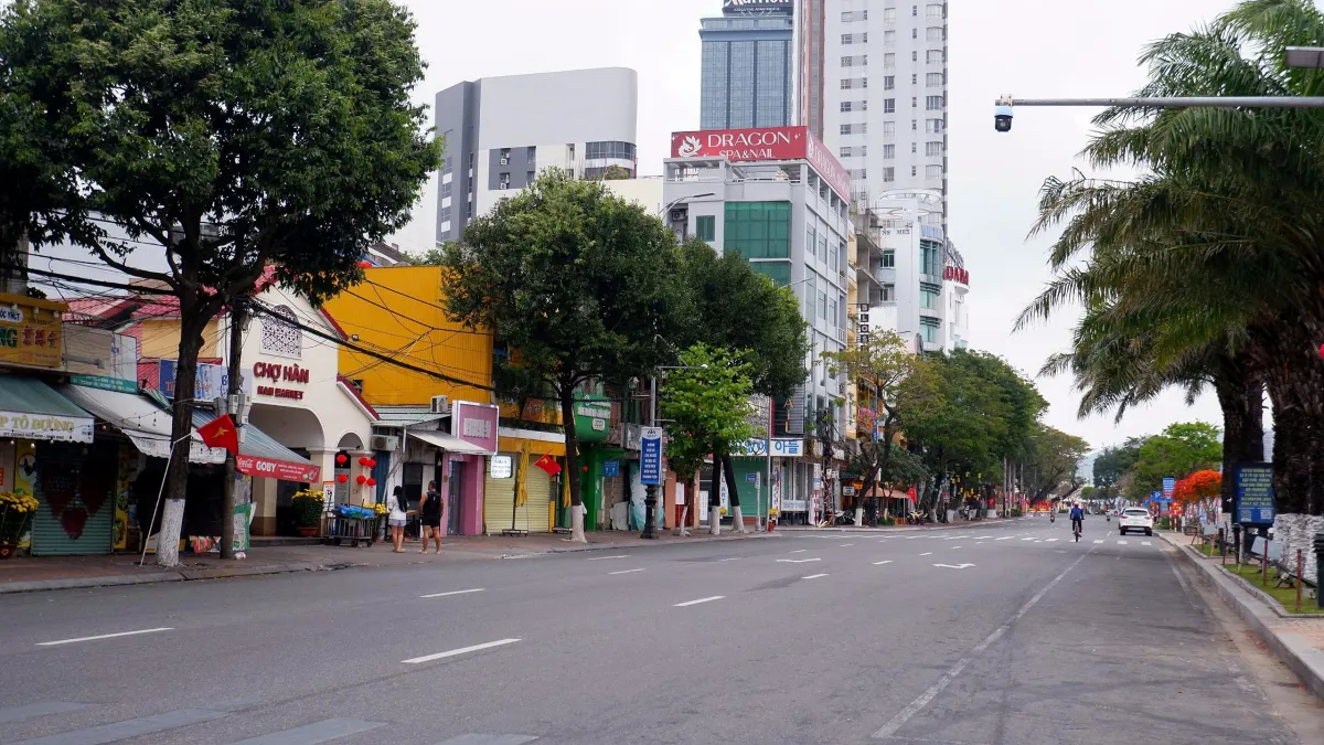 An almost empty Bach Dang Street with Han Market's cream gatehouse on the left, shops with red flags, high-rise buildings behind and palm trees along the river side