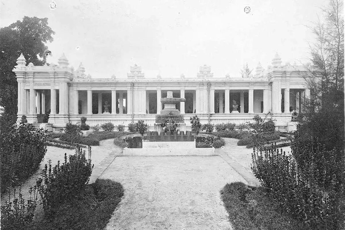 Black-and-white photo of a long colonnaded building with small tower-shaped ornaments on the roof, behind a formal garden