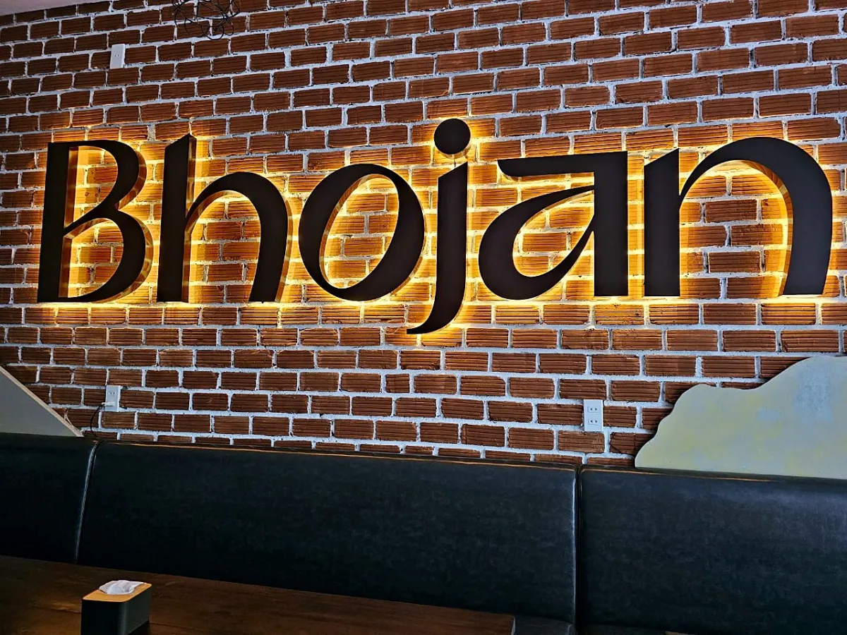 Black metal Bhojan sign lit in gold on a red brick wall above a long black leather bench seat