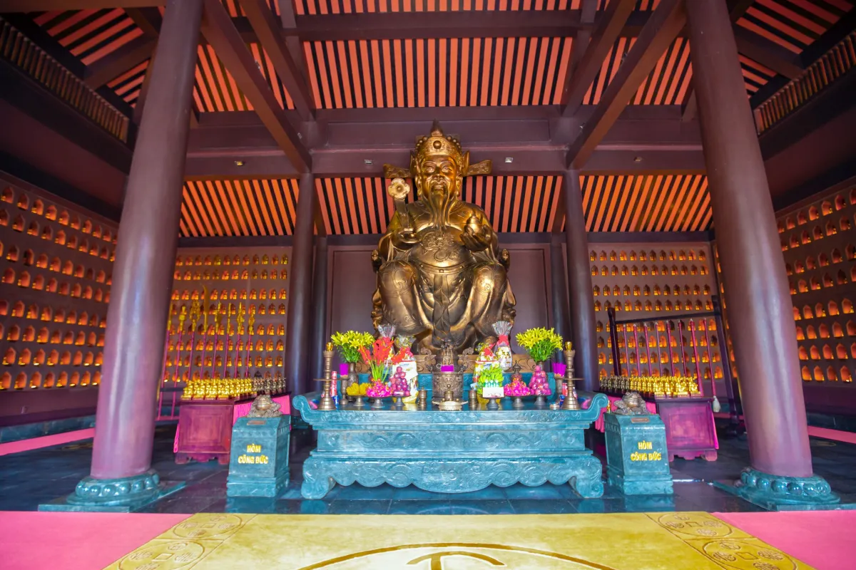 Bronze statue of the God of Fortune seated behind a carved stone altar with offerings, inside the Than Tai temple