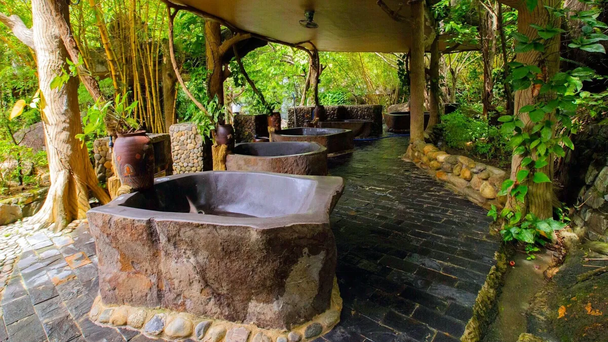Carved stone bathtubs in a row under a covered walkway among trees and bamboo at the mud-bath area