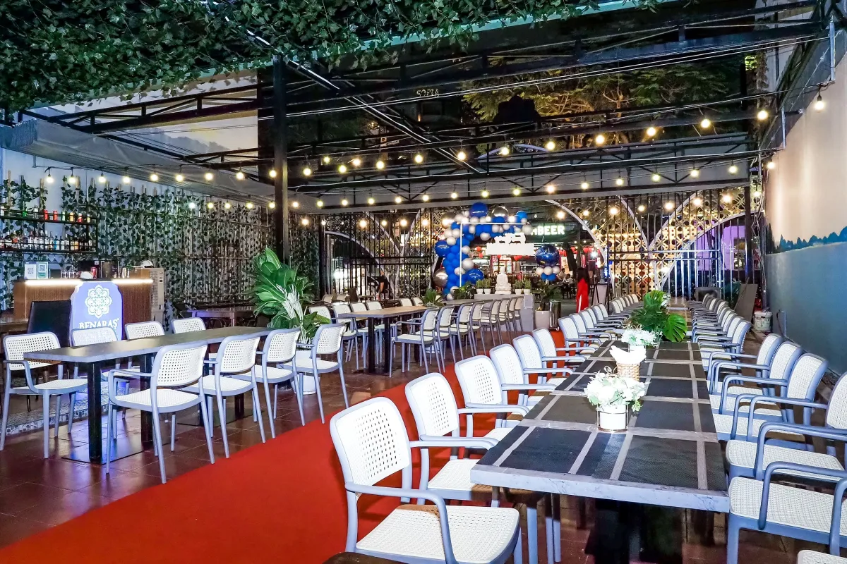 Covered courtyard at Benaras Central at night with a vine-covered roof, strings of round bulbs, long gray tables, white chairs and a red carpet aisle