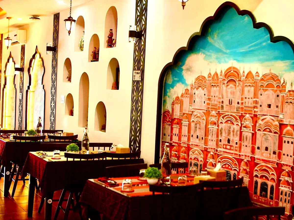 Dining room at Indian Aroma with a large painted mural of the Hawa Mahal in an arched frame, lit niches and tables with red and black checked cloths
