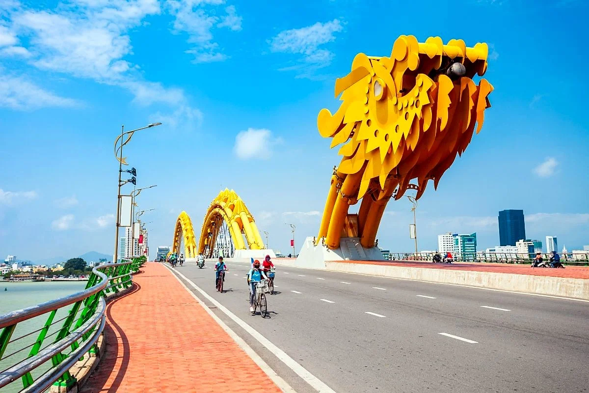 Dragon Bridge in daytime, with its yellow dragon head beside the road and a few people cycling along the road