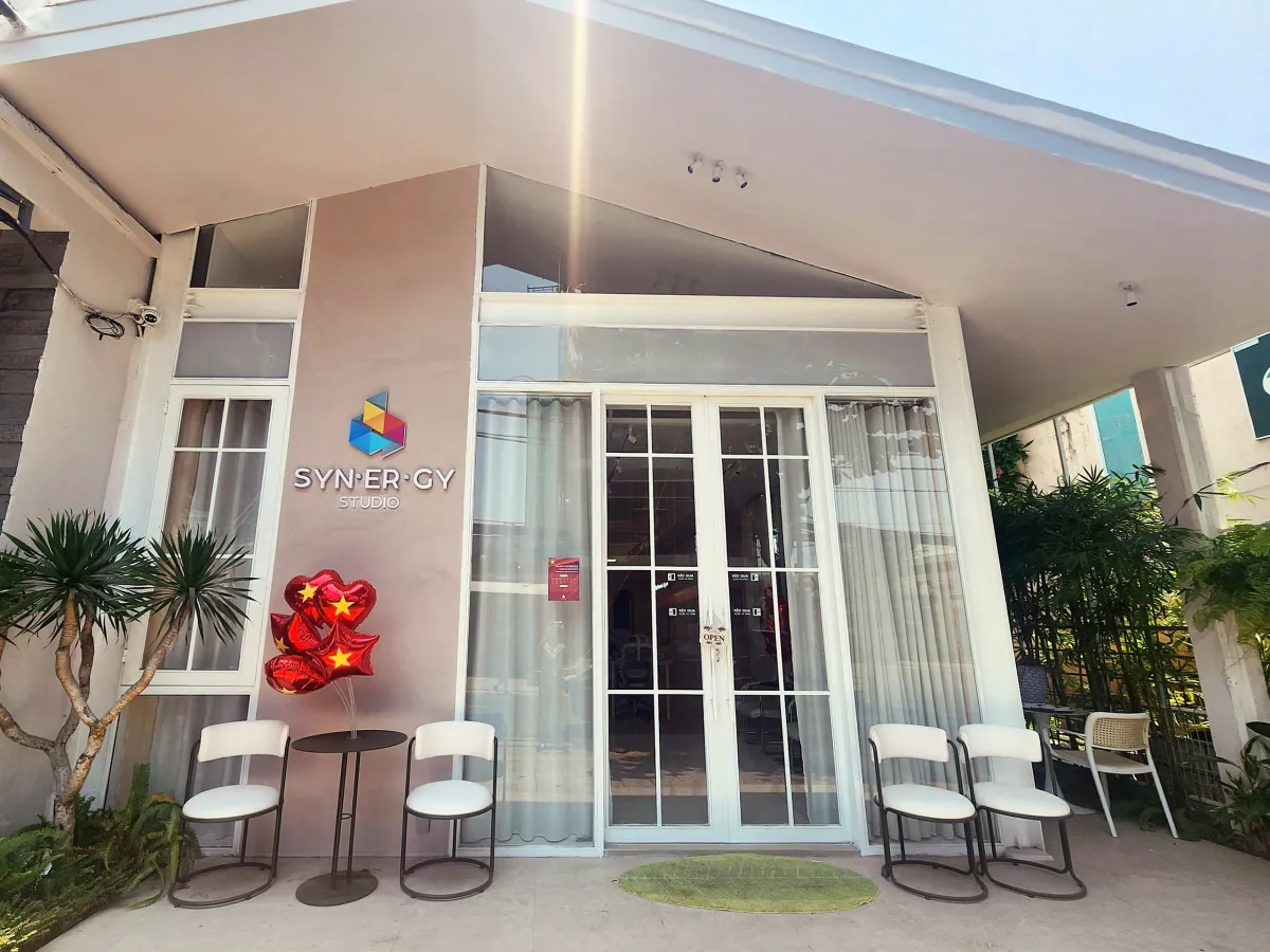 Front of Synergy Studio, a pale pink house with white-framed glass doors, the Synergy Studio sign and white chairs on the porch