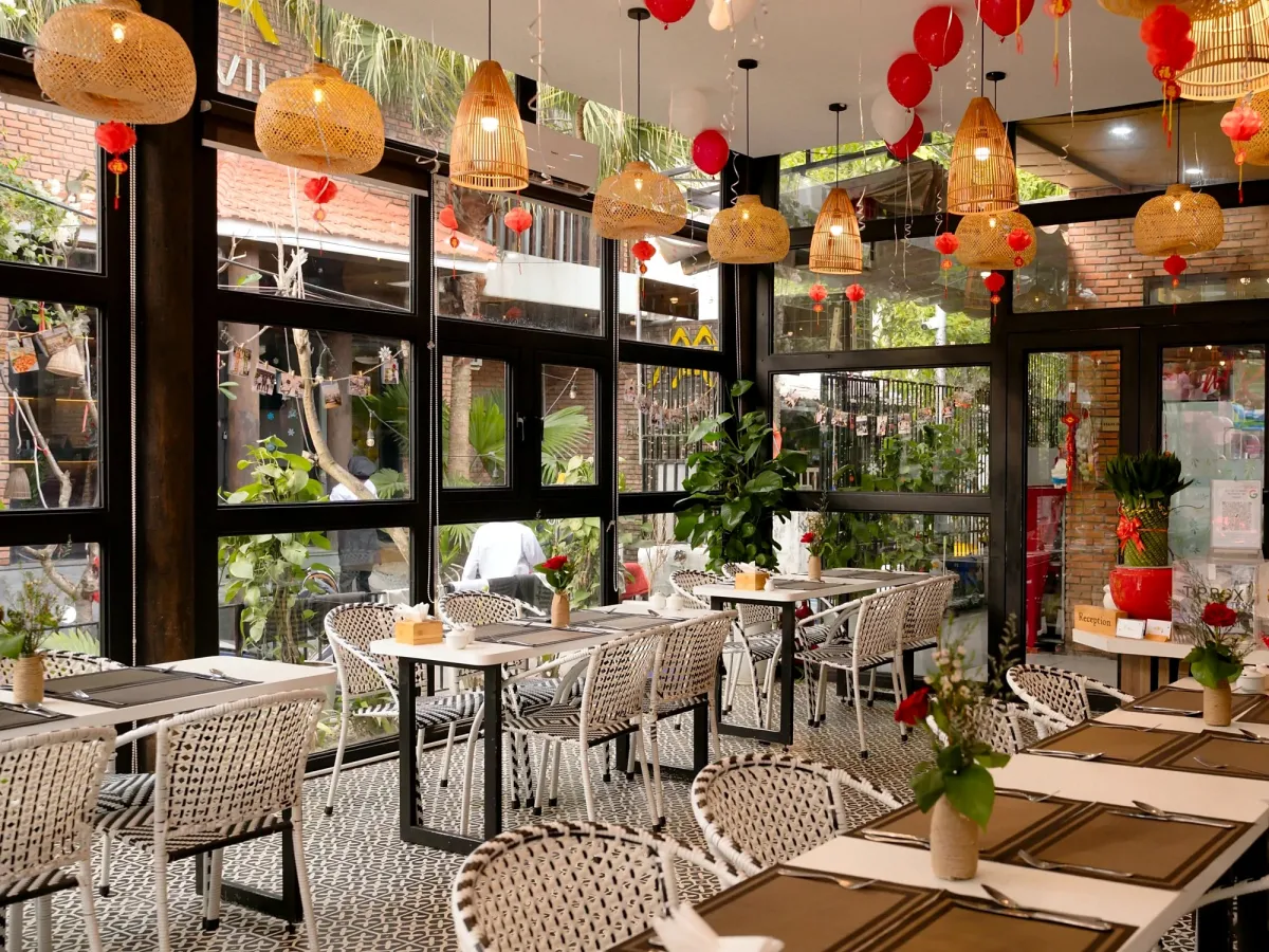 Glass-walled dining room at Manvar Rasoi with white and black woven chairs, red roses on the tables, rattan lamps and red lanterns