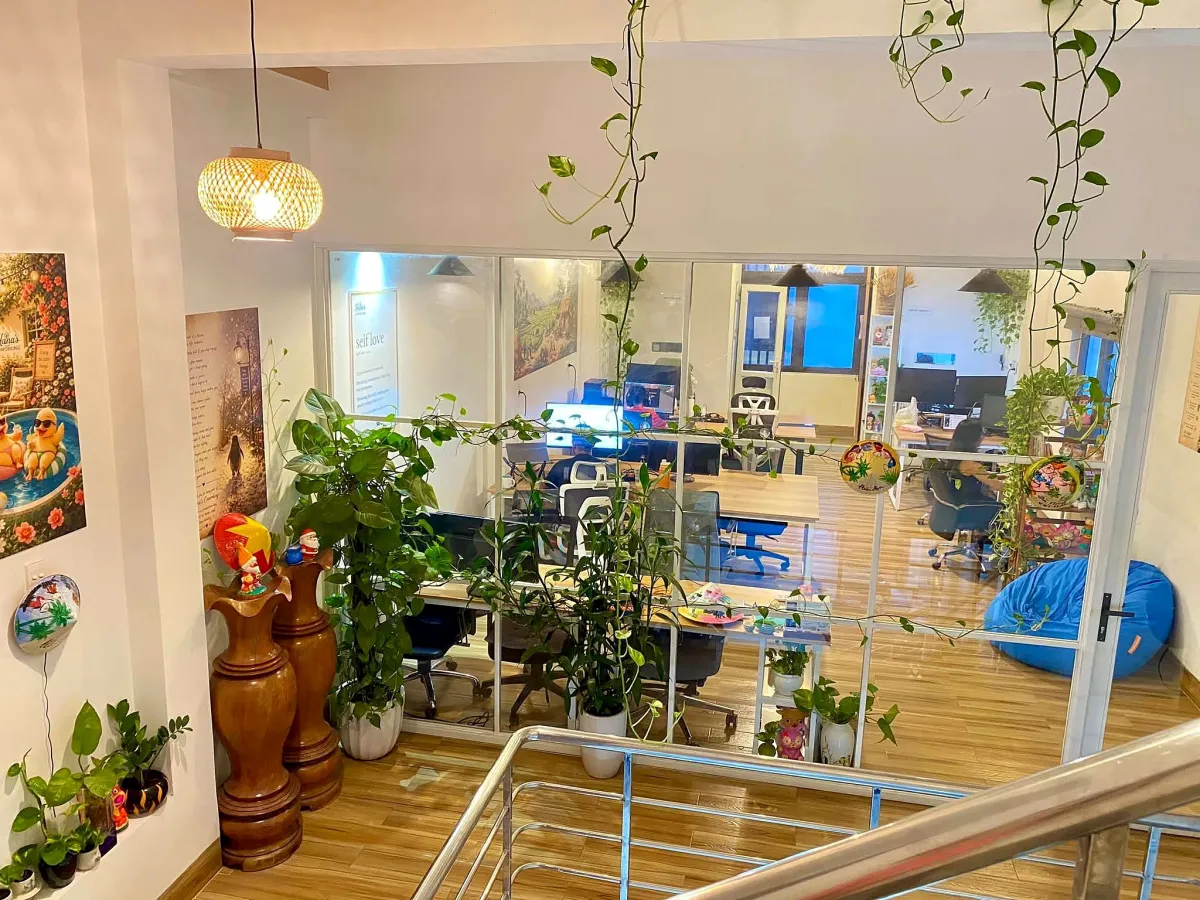 Glass-walled workroom at Hana's Coworking with wooden desks, white mesh chairs, monitors, a blue beanbag and trailing plants, seen from the stairs