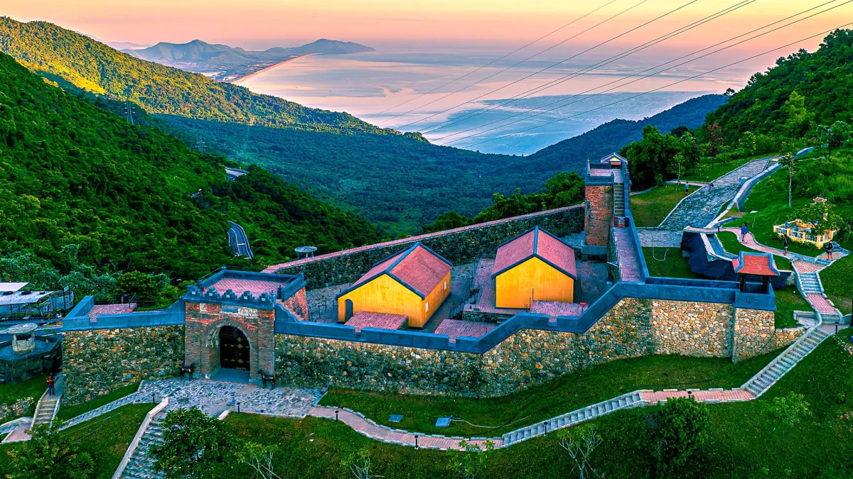 Hai Van Gate, an old stone gate and wall beside two tile-roofed buildings, seen from above in morning light, with Lang Co bay behind