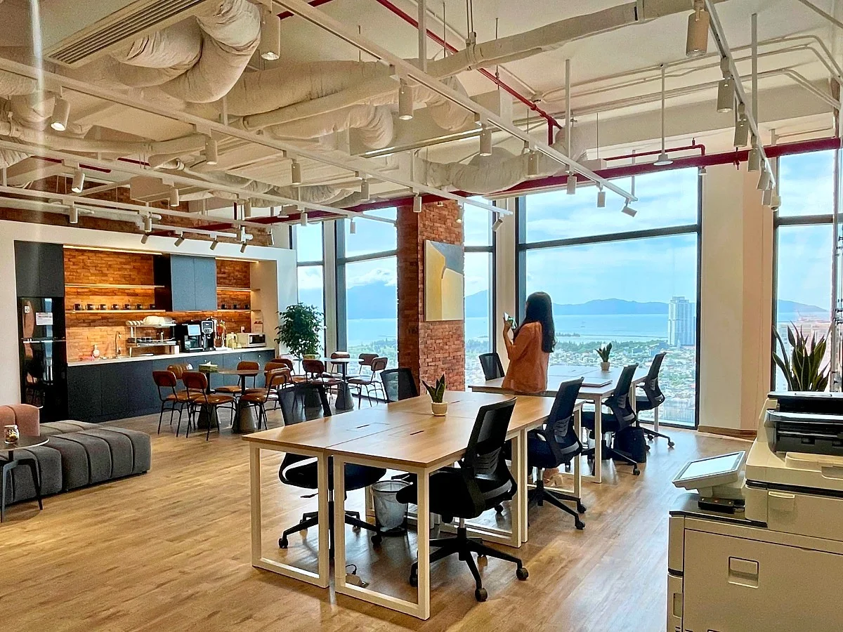 Long wooden desks and black office chairs on the 30th floor at cirCO Da Nang City Centre, with a coffee bar on the left and windows looking over the bay