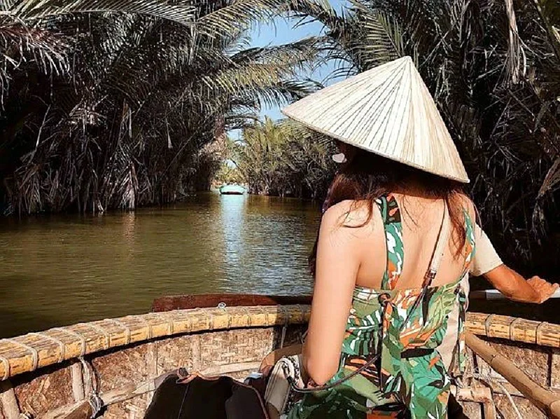 Seen from behind, a woman in a conical hat sits in a round basket boat on a calm channel lined with nipa palms