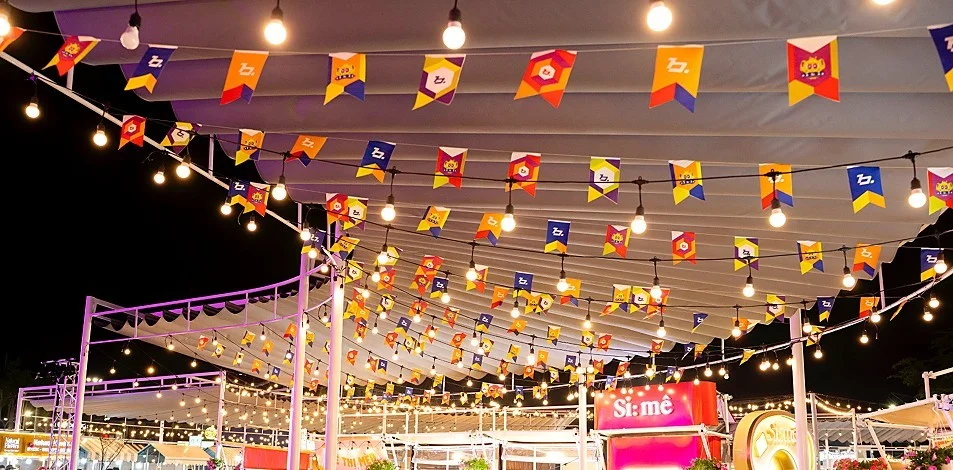 Strings of bulbs and colorful pennant flags hung under a white canopy over the B.Fair stalls at night, with a pink Si:mê sign below
