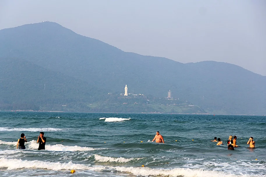Swimmers in the waves at My Khe, with the Goddess of Mercy statue and a pagoda tower on the Son Tra peninsula in the distance
