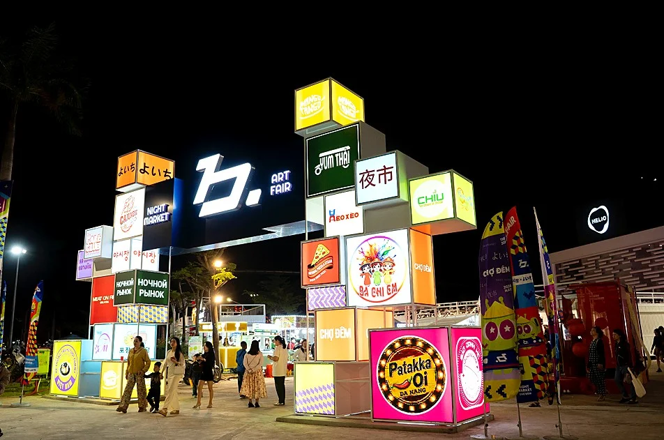 The B.Fair gate at night, with a large letter b and the words Art Fair, and stacked light boxes showing night market in several languages and stall names