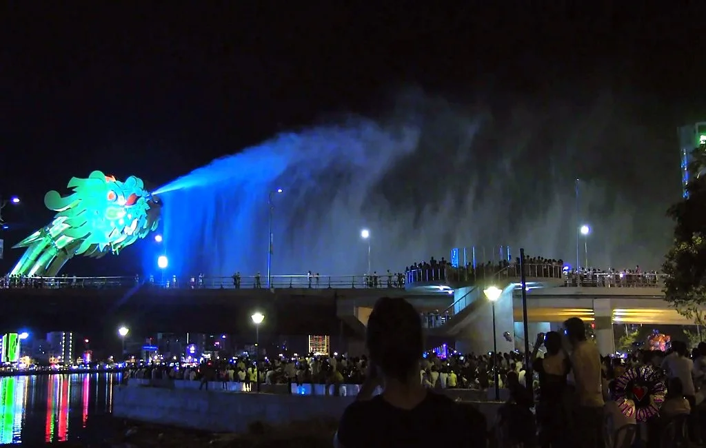 The dragon's head lit green, spraying a wide blue cloud of water over the bridge, with crowds watching from the bridge and the riverbank