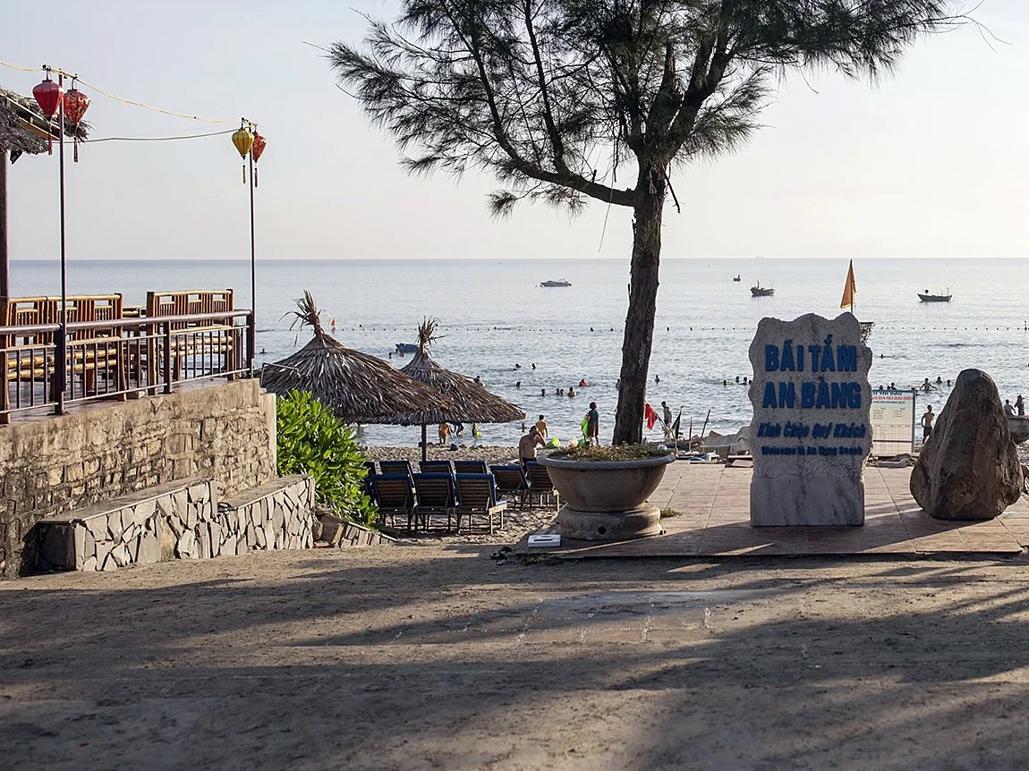 The entrance to An Bang beach with a white welcome stone reading Bai tam An Bang, thatched umbrellas, loungers and swimmers in a calm sea