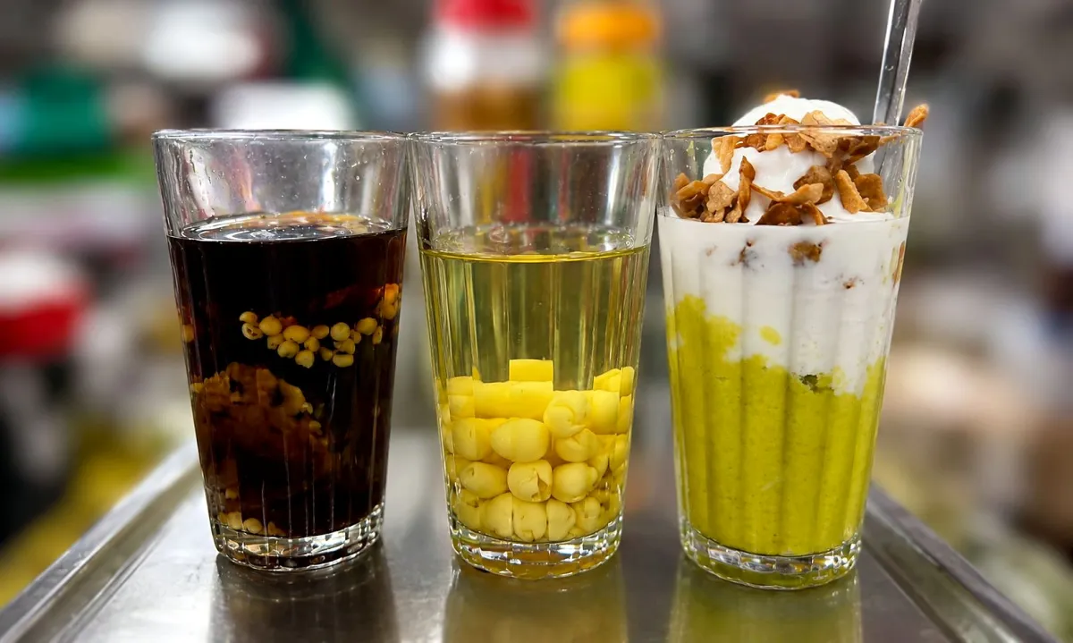 Three glasses on a steel counter: two sweet soups with beans and lotus seeds, and a layered avocado dessert topped with white cream and toasted coconut