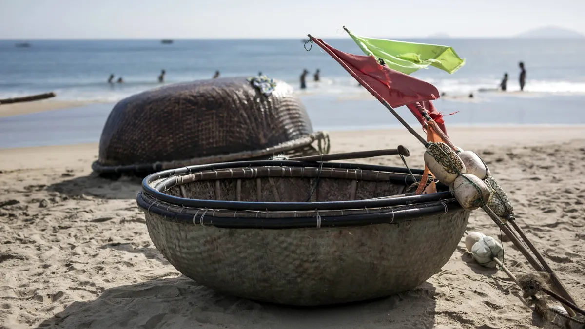 Two round bamboo basket boats on the sand at An Bang with red and green flags tied to a pole and swimmers in the sea behind