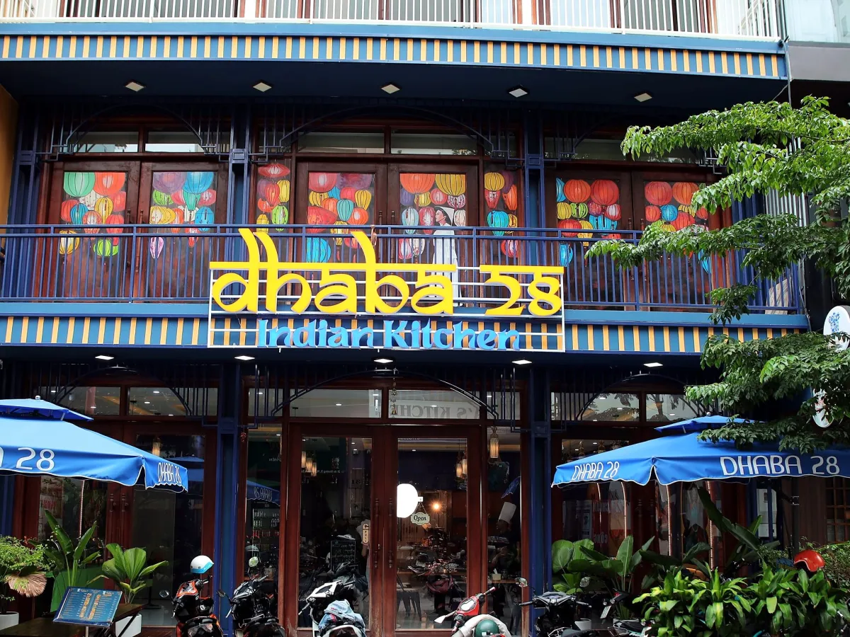 Two-story front of Dhaba 28 with a yellow sign, lantern paintings on the upstairs windows, a striped awning and blue umbrellas