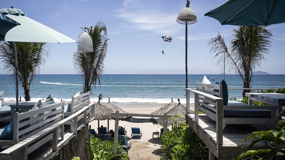 View from a white wooden beach deck with cushioned benches and umbrellas, past a sign reading The Deck House, to loungers on the sand and a parasail over the sea